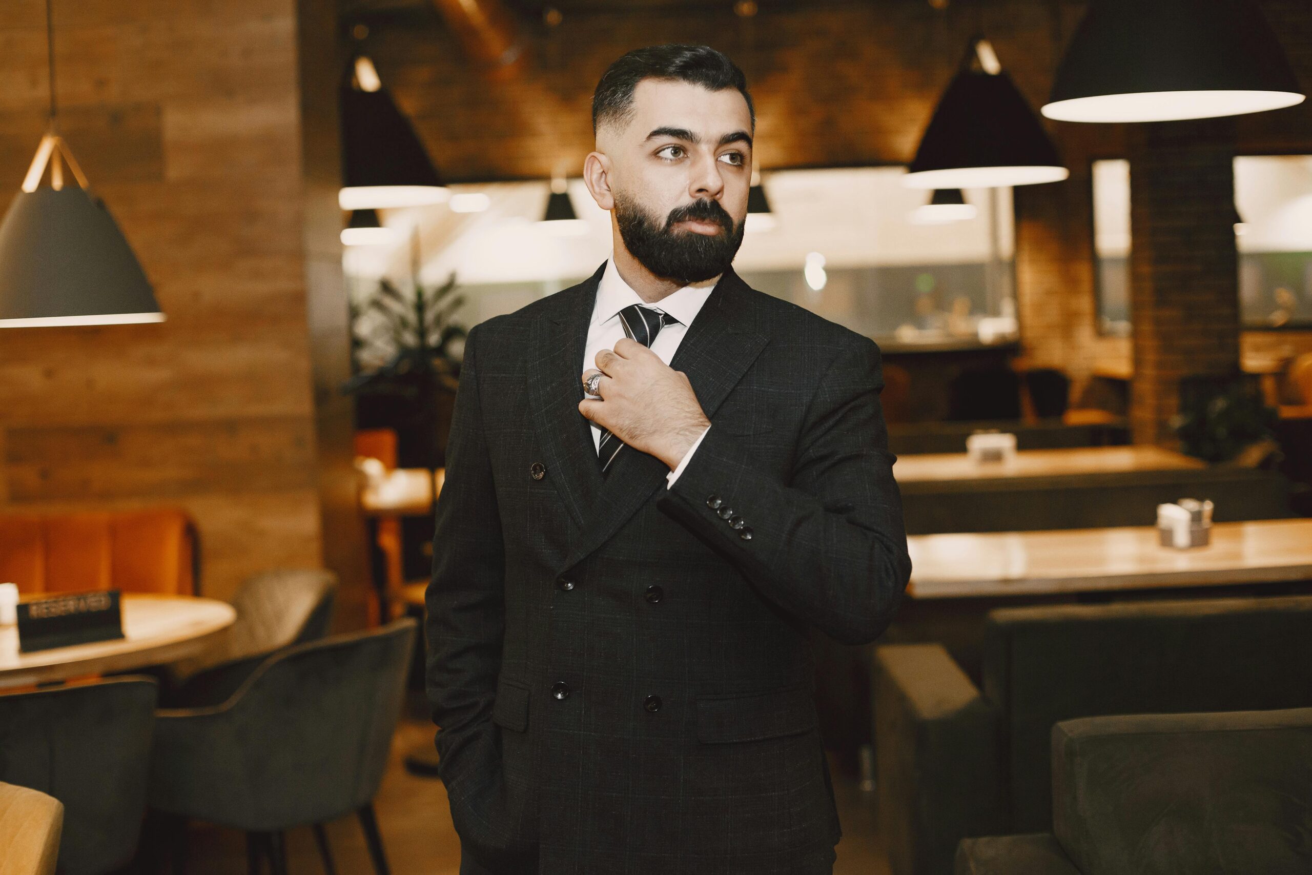 Stylish man in a black suit and tie standing confidently in a modern restaurant setting.
